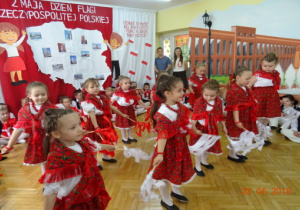 Na tle dekoracji z mapą Polski i dziećmi z chorągiewkami tańczą dziewczynki przebrane za góralki.